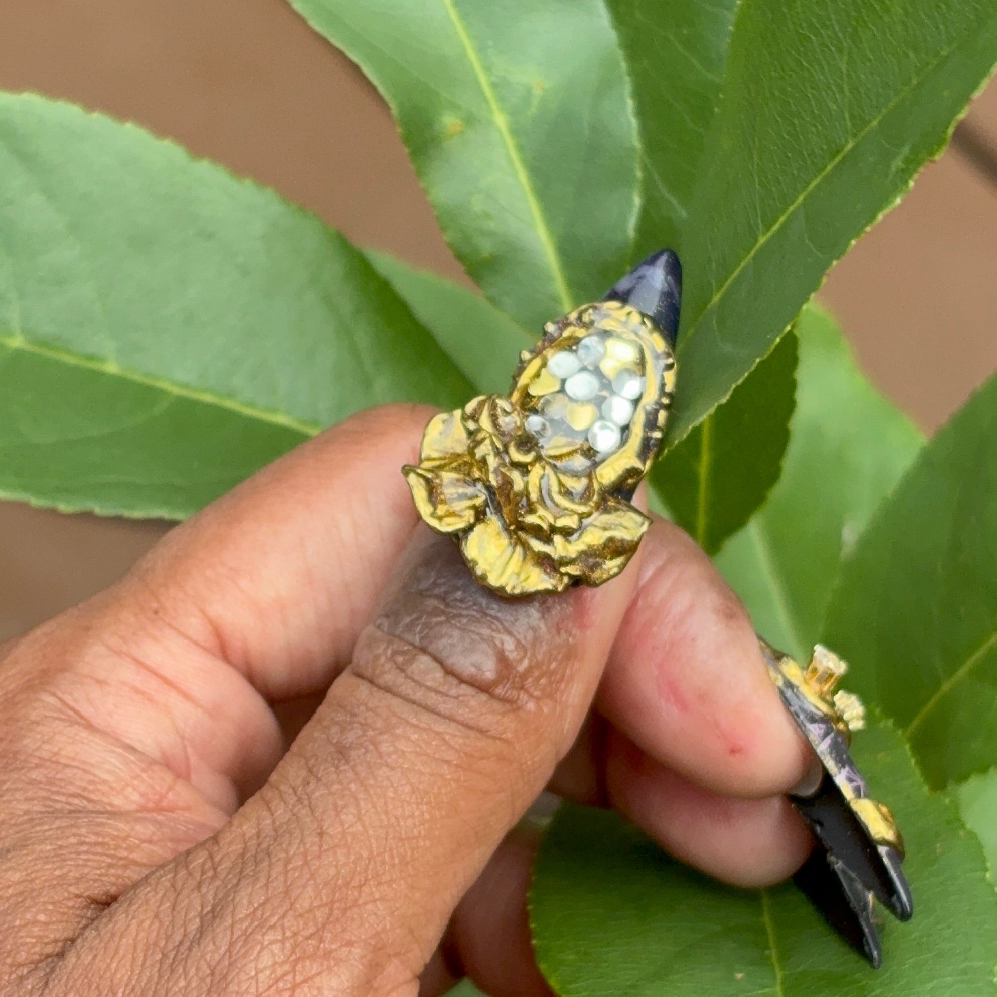 Hand showing thumb with press ons against dark skin with a dark base and chrome and 3D work similar to jewelry.