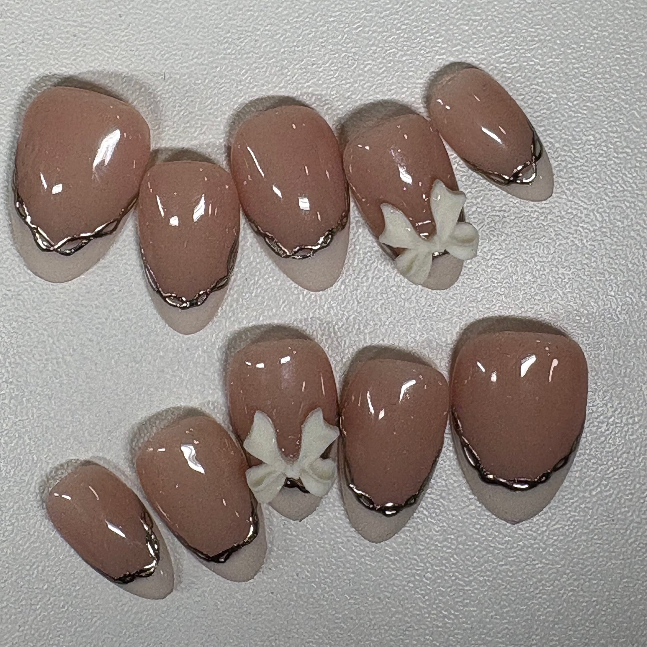 Each nail features a chrome metallic border that defines the edge, similar to the binding of a satin shoe. We’ve added a single 3D white bow on the ring finger as a focal point.