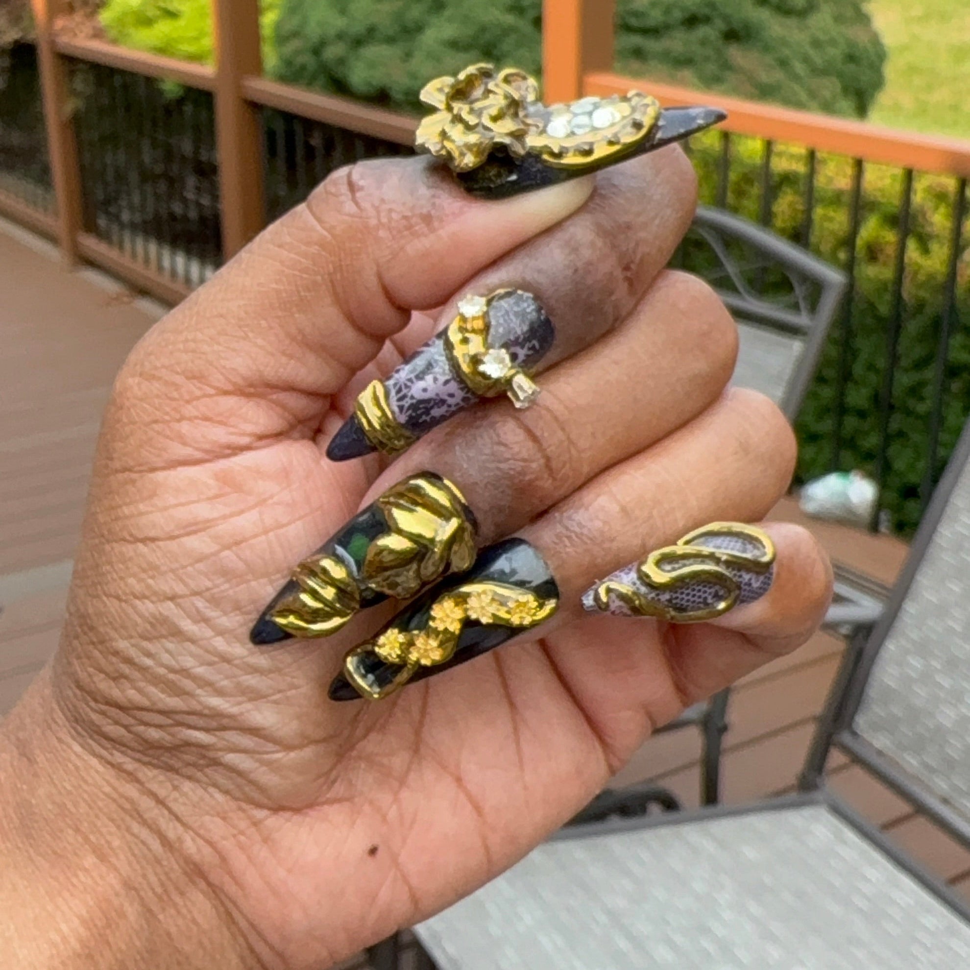 Hand showing 5 different press ons against dark skin with dark base and chrome and 3D work similar to jewelry.