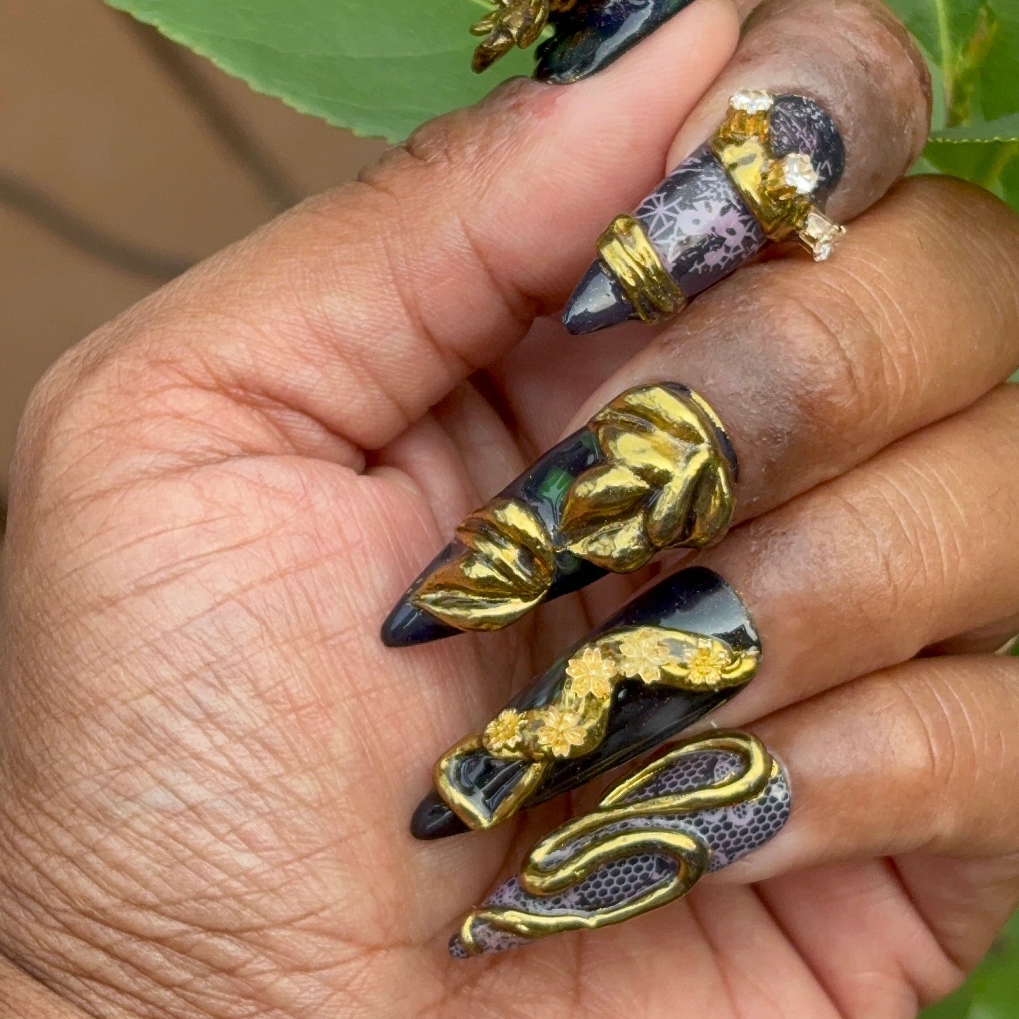 Hand showing press ons against dark skin with 3D work with gold chrome and charms looking like jewelry nails