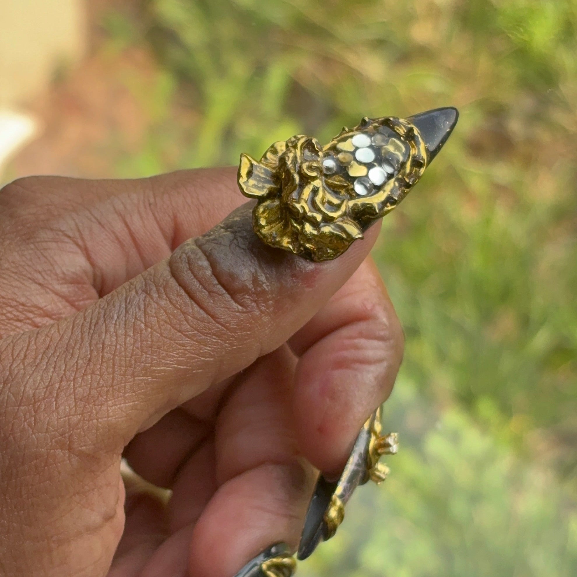 A hand with a luxury press on against dark skin showing medium almond nails with jewelry level gold chrome and charms.